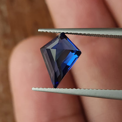 Tilted blue kite sapphire in tweezers in front of a hand, showing dark crown reflections and its pointed outline.