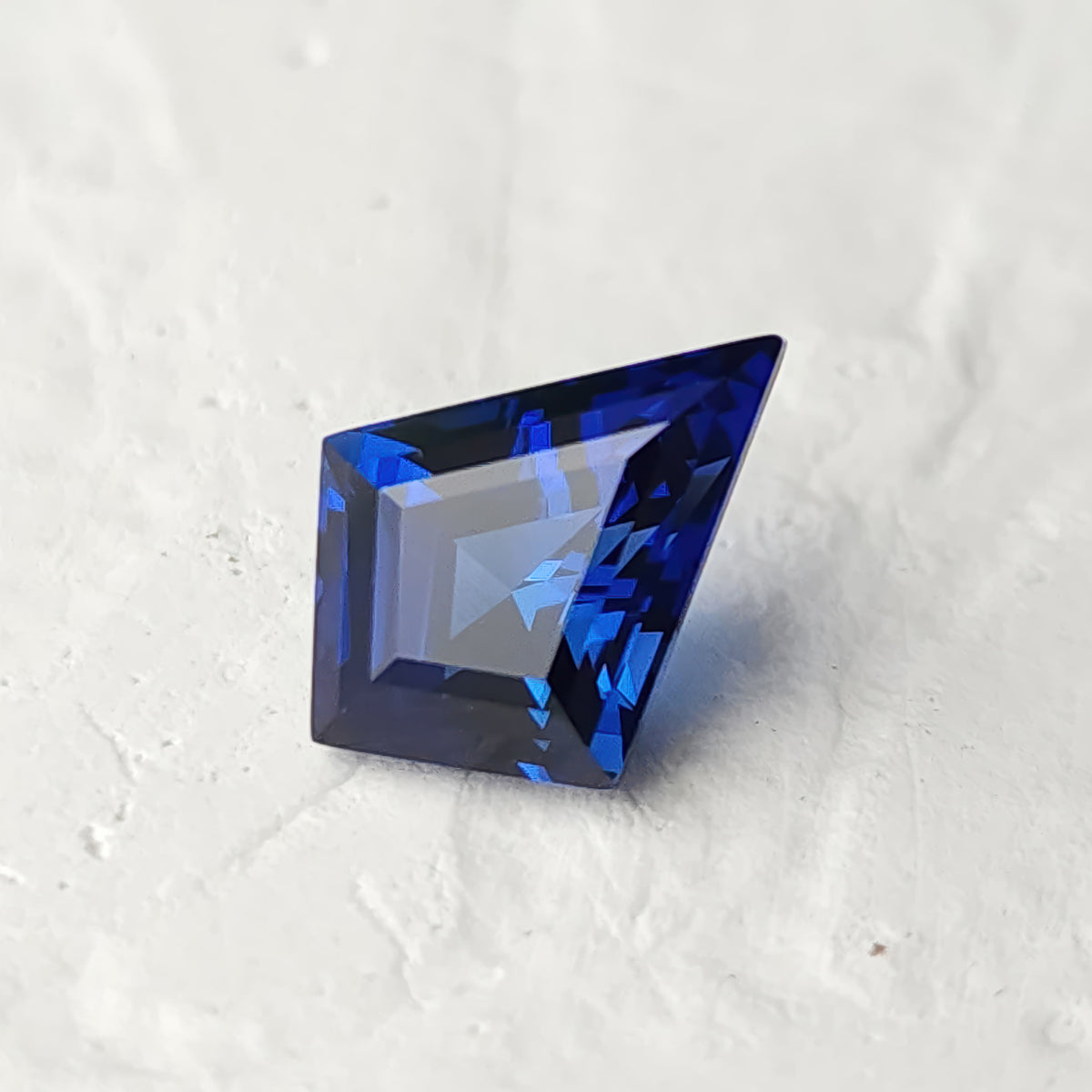 Blue lab-grown kite sapphire resting on a white surface, with broad pale reflections across its geometric crown.