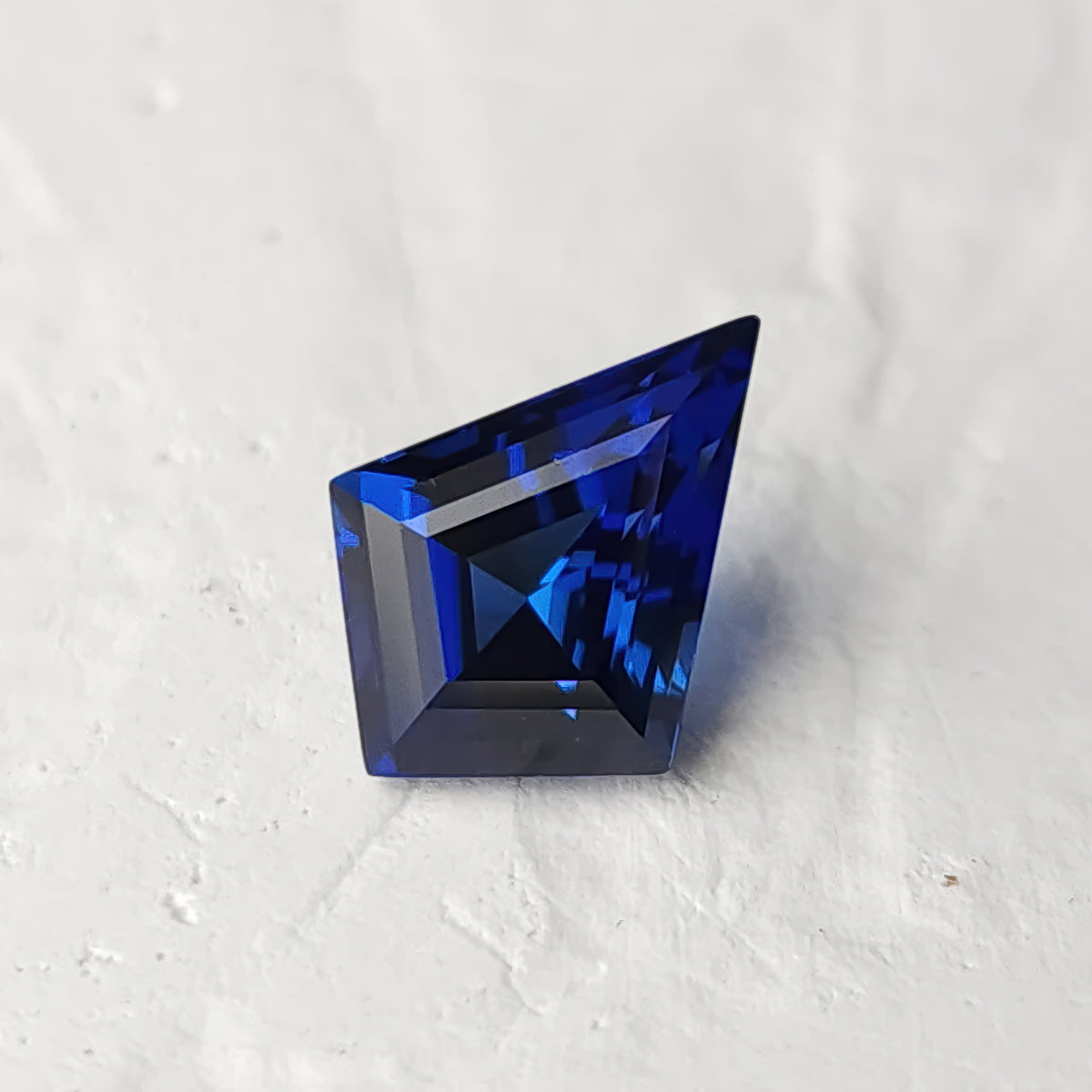 Royal blue kite-cut sapphire on a light textured surface, showing dark central facets and bright blue reflected bands.