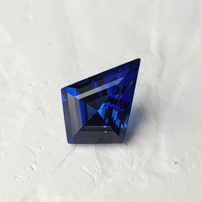 Royal blue kite-cut sapphire on a light textured surface, showing dark central facets and bright blue reflected bands.