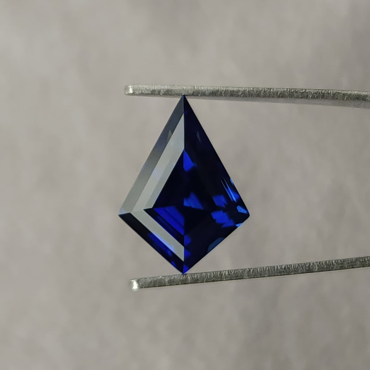 Kite sapphire held upright in tweezers against a gray background, with blue reflections and a pale band along the edge.
