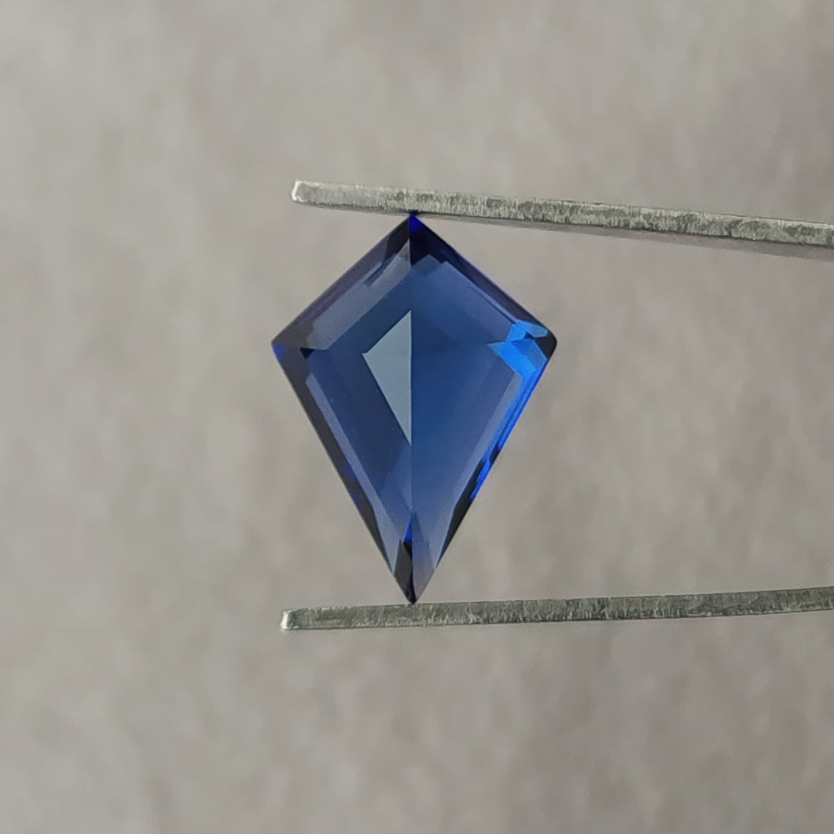 Reverse view of the blue kite sapphire held in tweezers, showing broad pale blue pavilion facets and its long point.