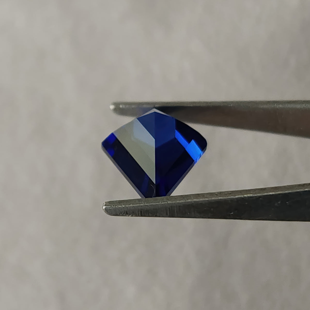 Angled pavilion of the royal blue kite sapphire in tweezers, with broad blue and pale reflected planes.