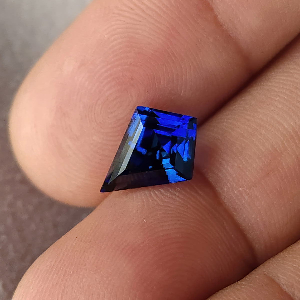 Angled kite-cut sapphire resting between fingertips, with dark center reflections and vivid blue edge flashes.