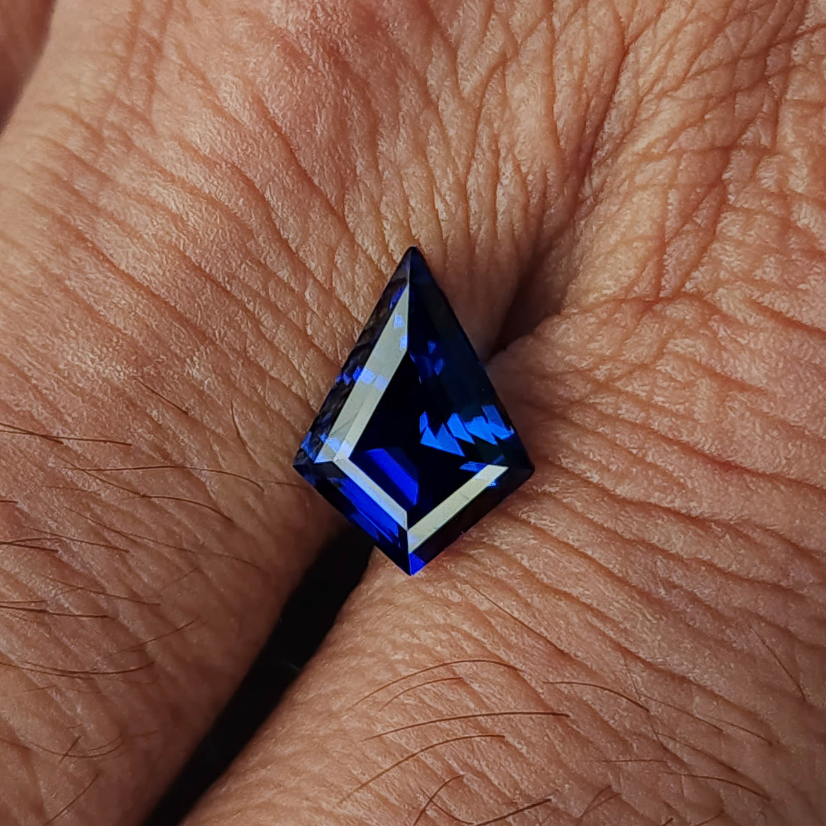 Royal blue kite sapphire on the back of a hand, with a dark supporting tool beneath it and skin texture for scale.