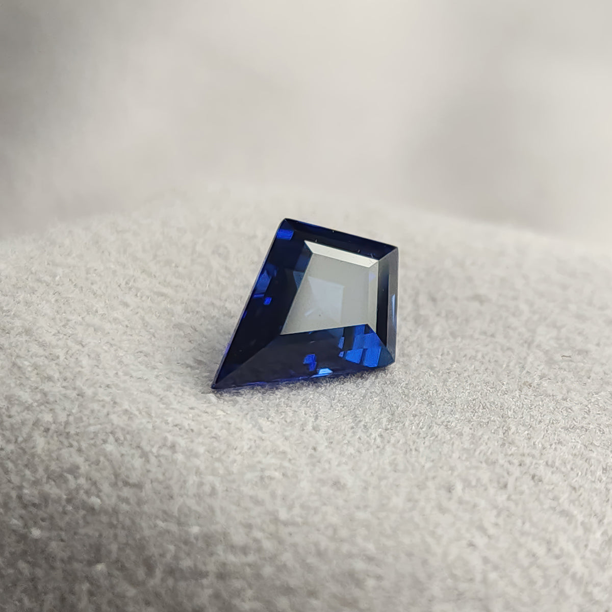 Kite sapphire leaning on a gray textured surface, with a broad pale face-up reflection and deep blue edges.