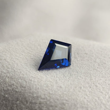 Kite sapphire leaning on a gray textured surface, with a broad pale face-up reflection and deep blue edges.