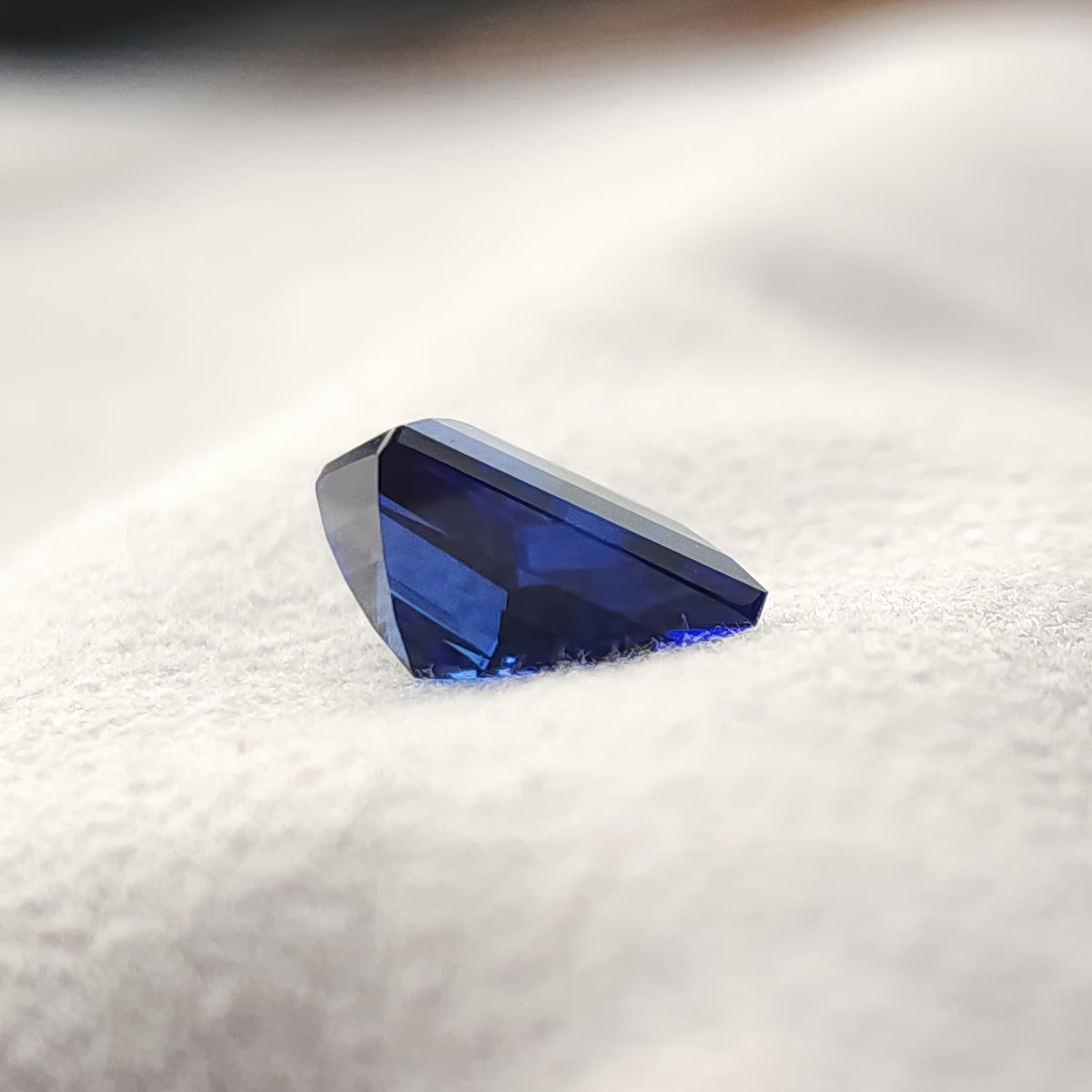 Low side view of the blue lab-grown kite sapphire on a light surface, showing its thickness and pointed profile.