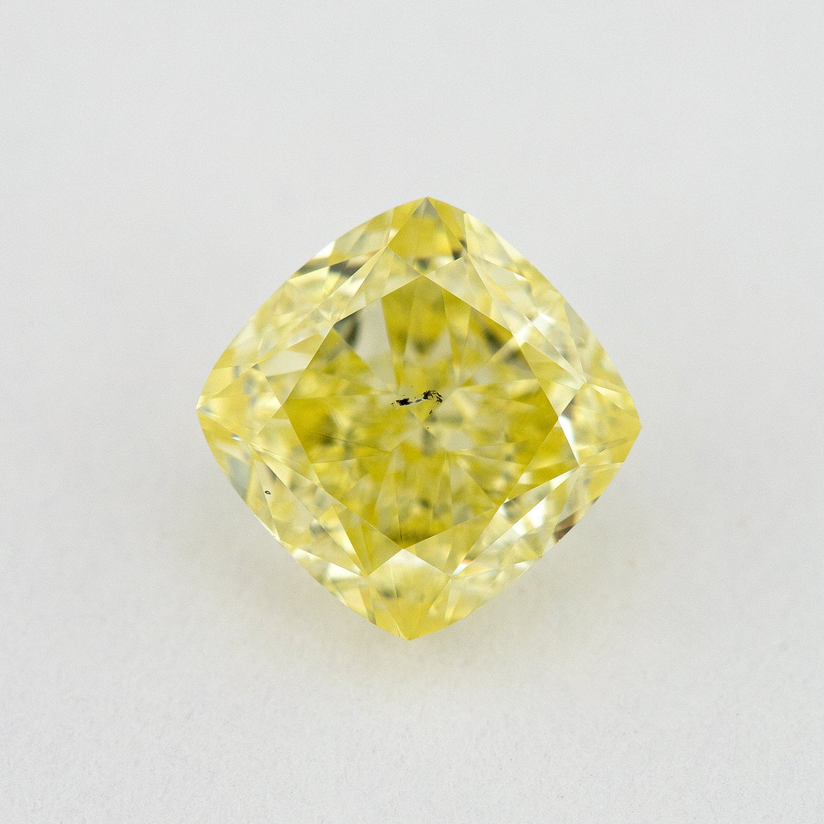 Face-up cushion-shaped yellow diamond on a pale surface, with small dark marks visible near the centre.