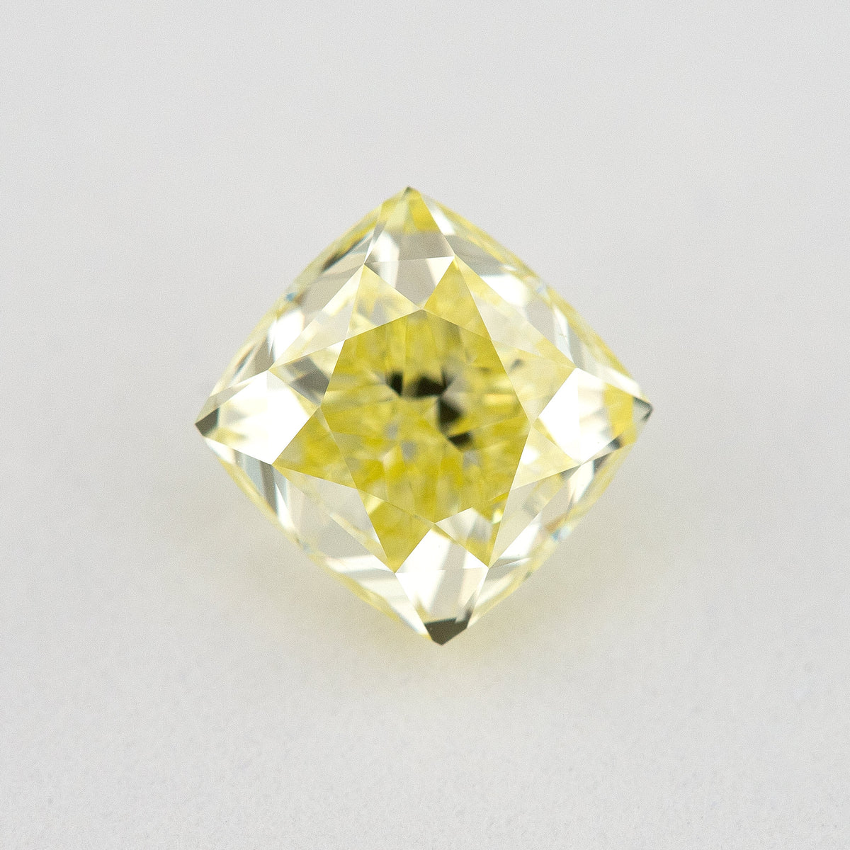 Face-up cushion-shaped yellow diamond on a pale surface, showing bright crown facets and dark central reflections.