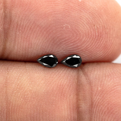 Black pear-shaped diamond pair resting on fingertips, with both faces and their small scale visible.