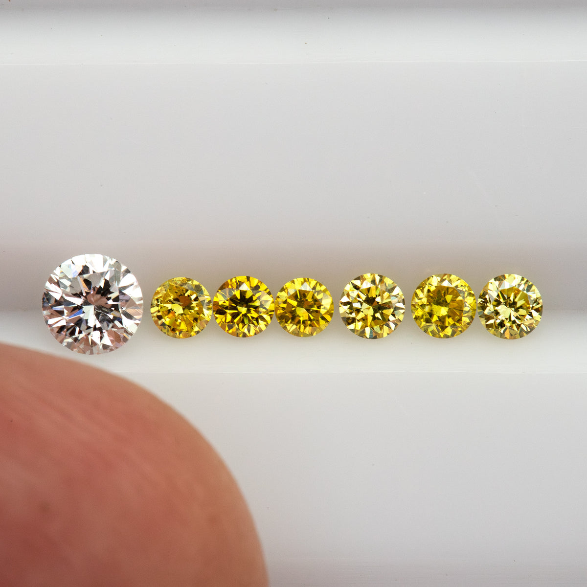 Six yellow diamonds in a face-up row with a fingertip for scale; the larger white comparison diamond is not included.