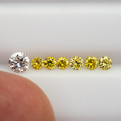 Six yellow diamonds in a face-up row with a fingertip for scale; the larger white comparison diamond is not included.
