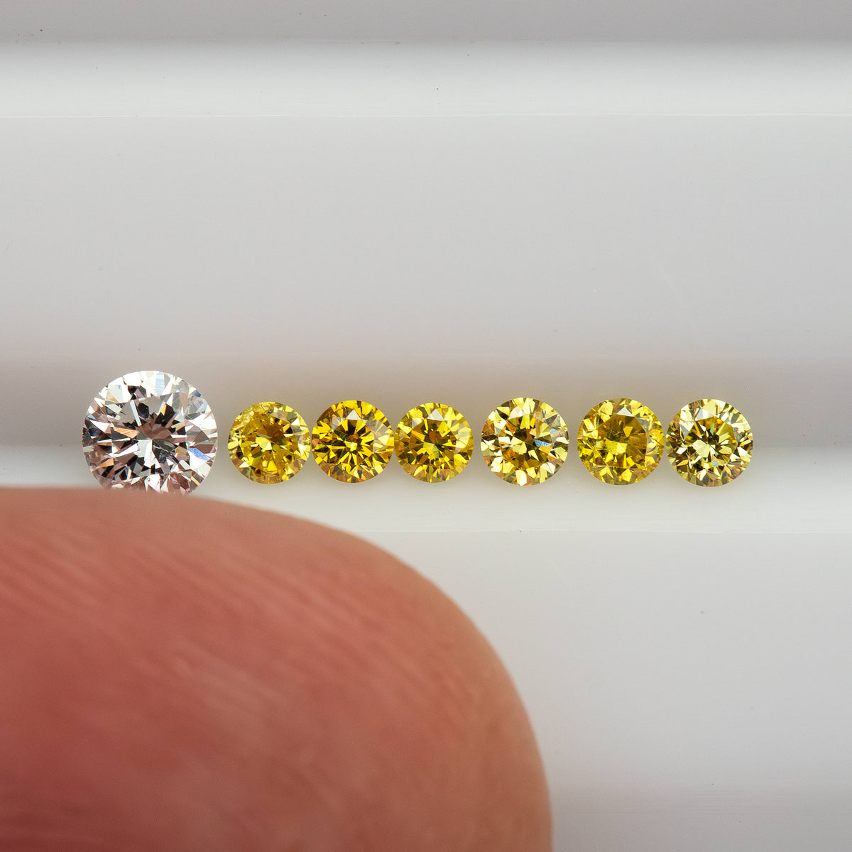 Lower-angle row of six yellow diamonds beside a fingertip; the white comparison diamond is not part of the parcel.