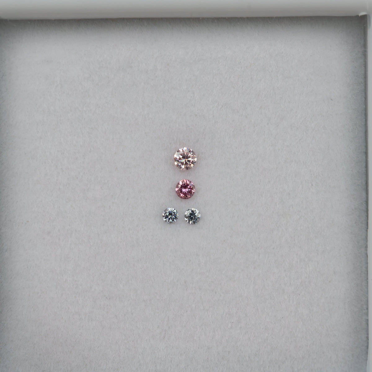 Wider face-up view of the four loose round diamonds centered on white padding, with two pink-looking stones over two smaller cool-toned stones.