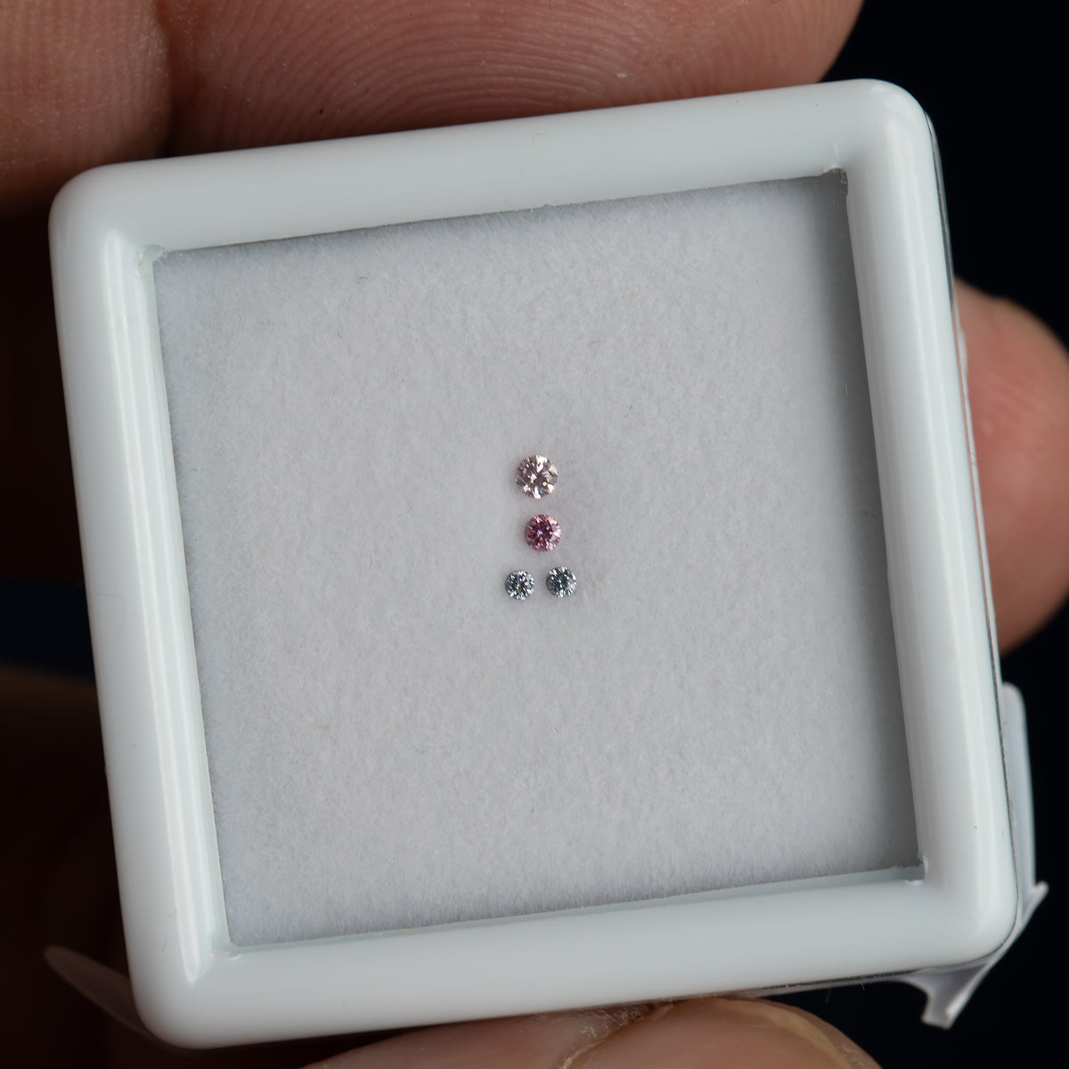 Hand-held white display frame containing four small round diamonds, with fingers around the frame and the entire set viewed face-up.
