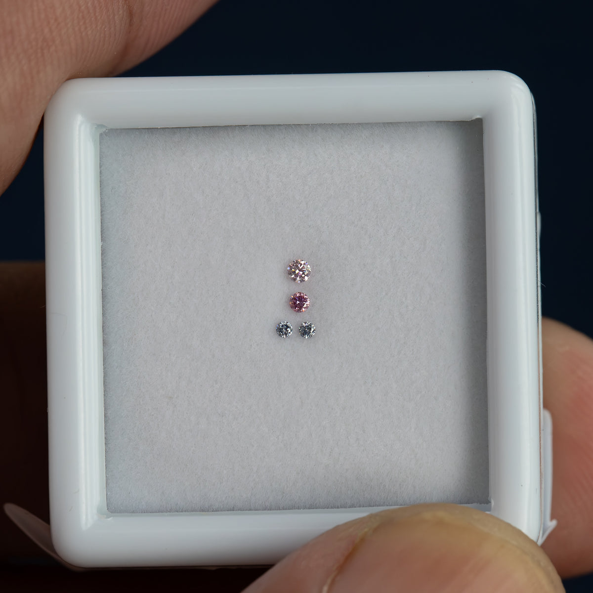 Four small round diamonds face-up in a white display frame held between fingers, showing their small scale in a hand-held view.
