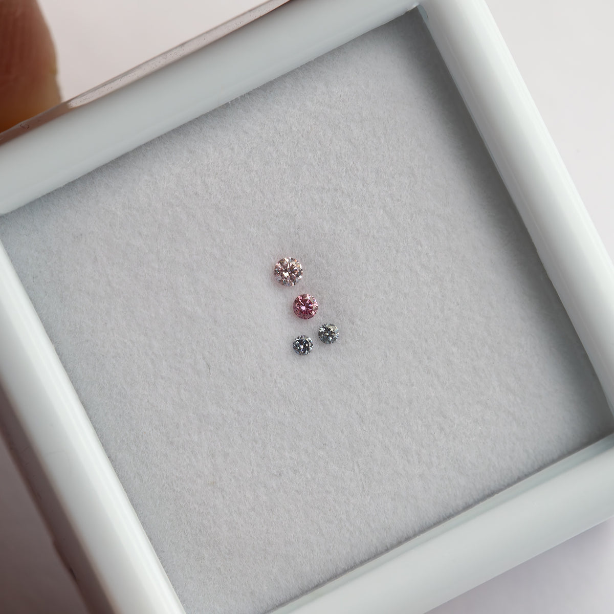 Four round diamonds resting face-up in a tilted white display frame, with two pink-looking stones above a pair of smaller blue-gray stones.