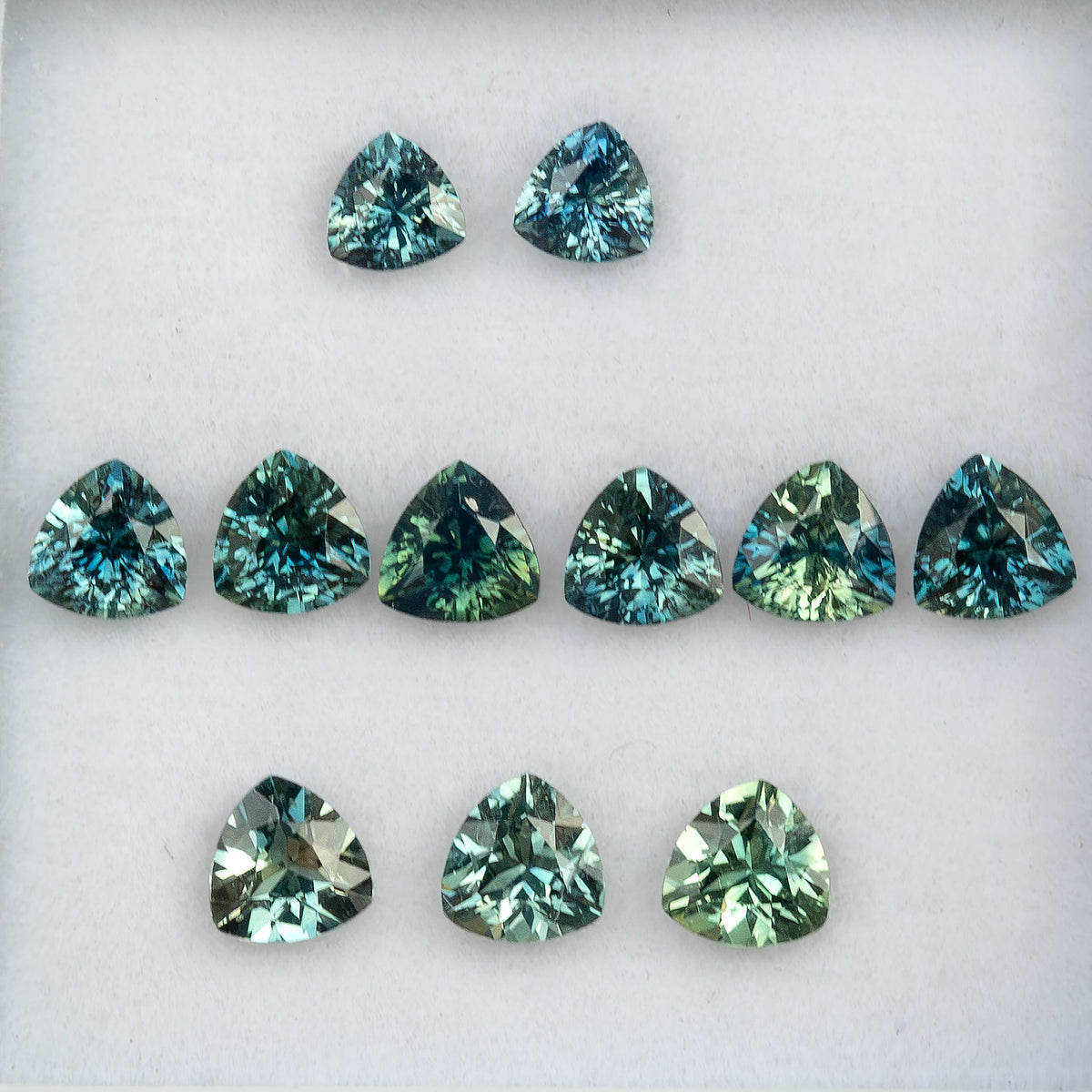 Face-up arrangement of natural teal trillion sapphires in this finite lot, showing blue-green to green color variation; stone #8 is sold.
