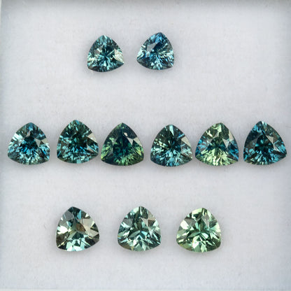 Face-up arrangement of natural teal trillion sapphires in this finite lot, showing blue-green to green color variation; stone #8 is sold.