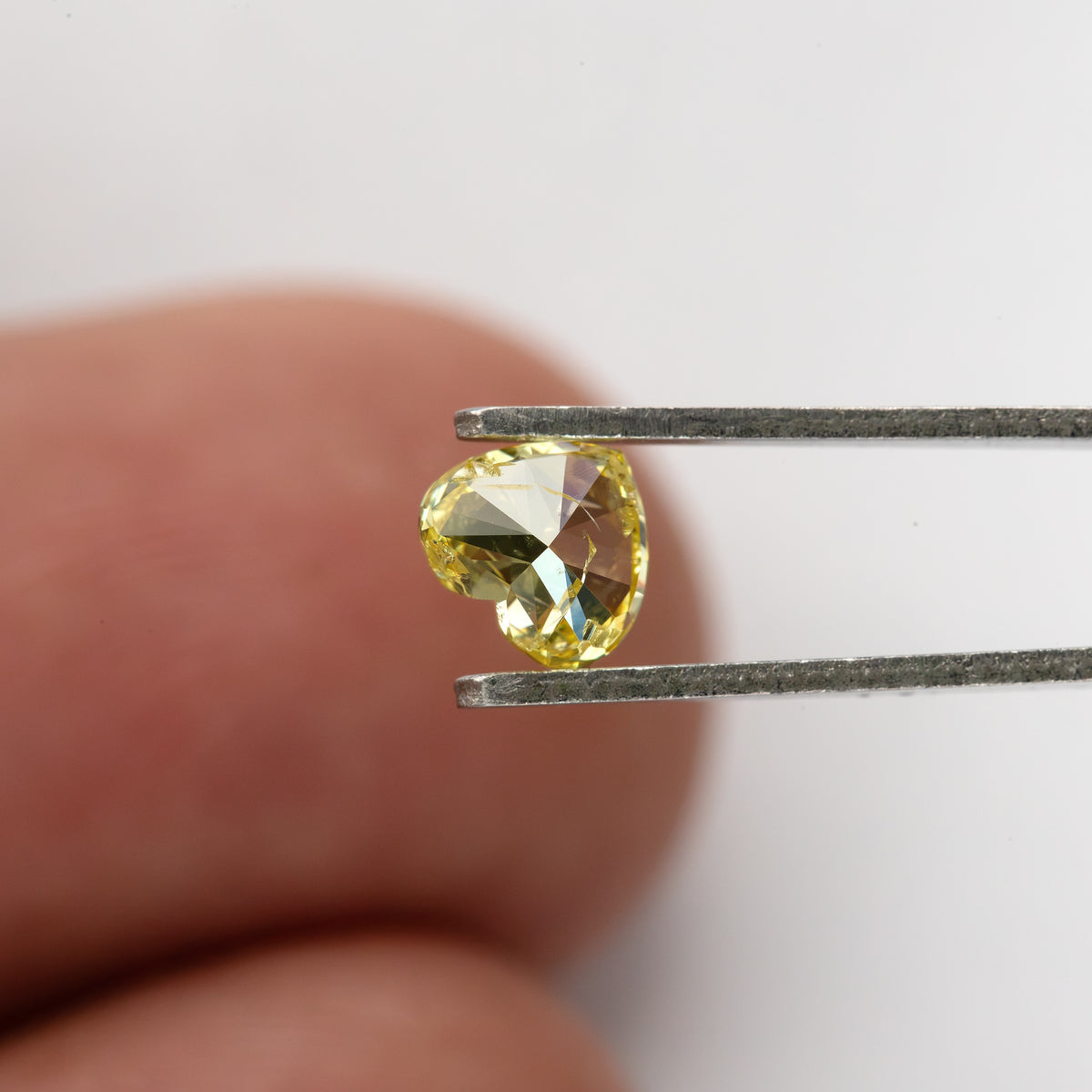 Yellow heart-shaped diamond turned back toward the camera in tweezers, with a fingertip behind it for scale.
