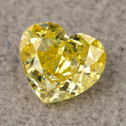 Yellow heart-shaped diamond resting face-up on a textured grey surface, with bright facet reflections and visible inclusions.