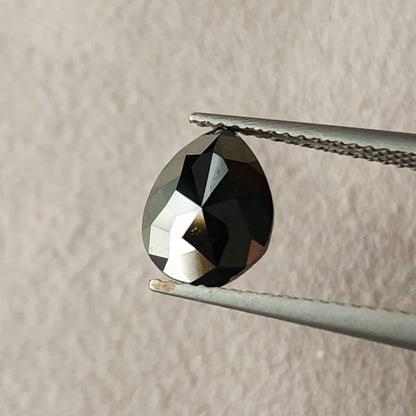 Second face-up angle of the pear rose-cut black diamond held point-up in tweezers, with a broad dark center and silver-toned reflections across the left facets.