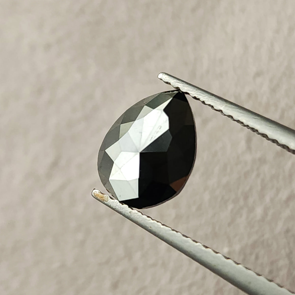 Face-up macro of the exact 1.63 carat pear rose-cut black diamond held in tweezers, showing its pointed outline, opaque black body and bright facet reflections.