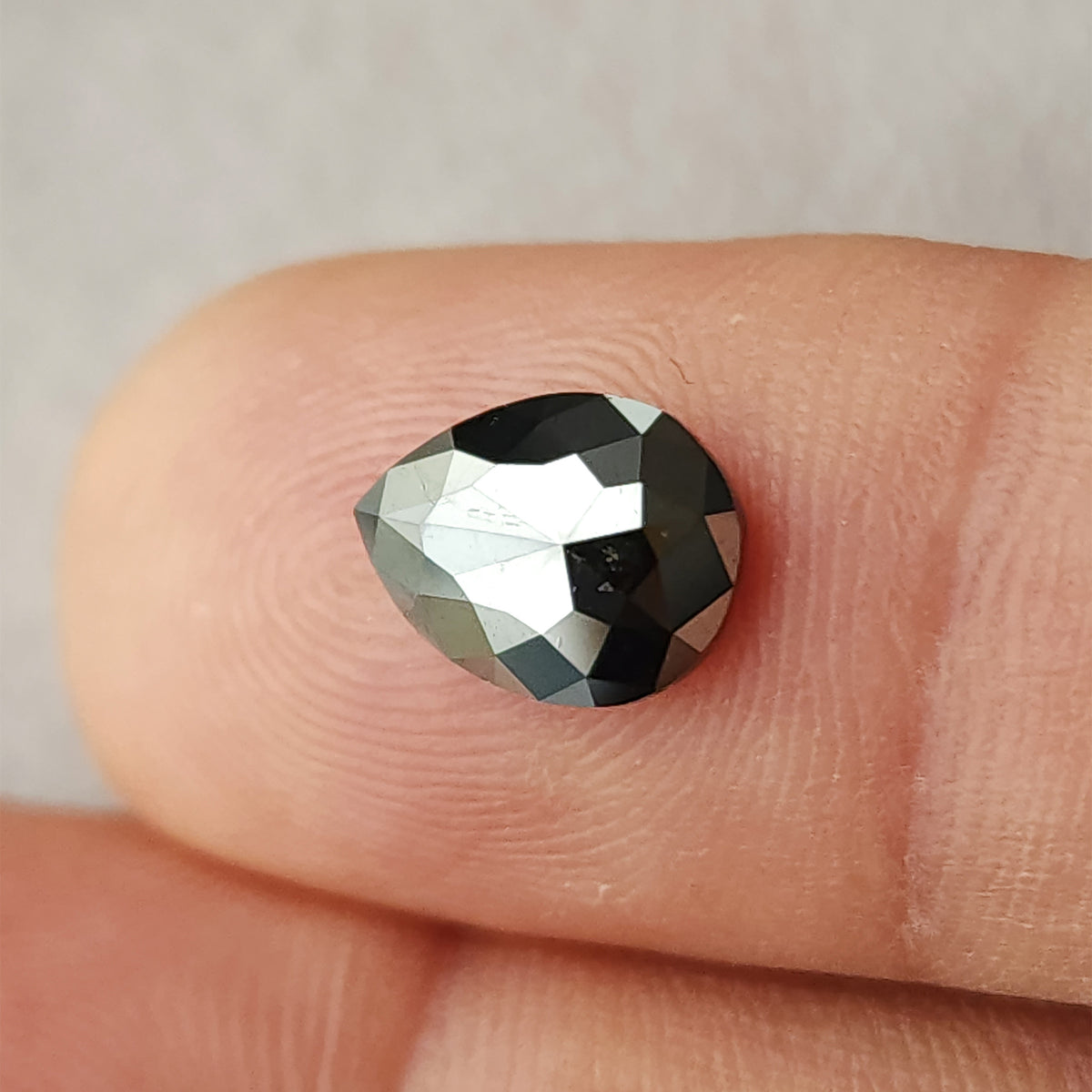 Angled view of the pear rose-cut black diamond resting on a fingertip, showing the rounded end, pointed tip and changing facet reflections.