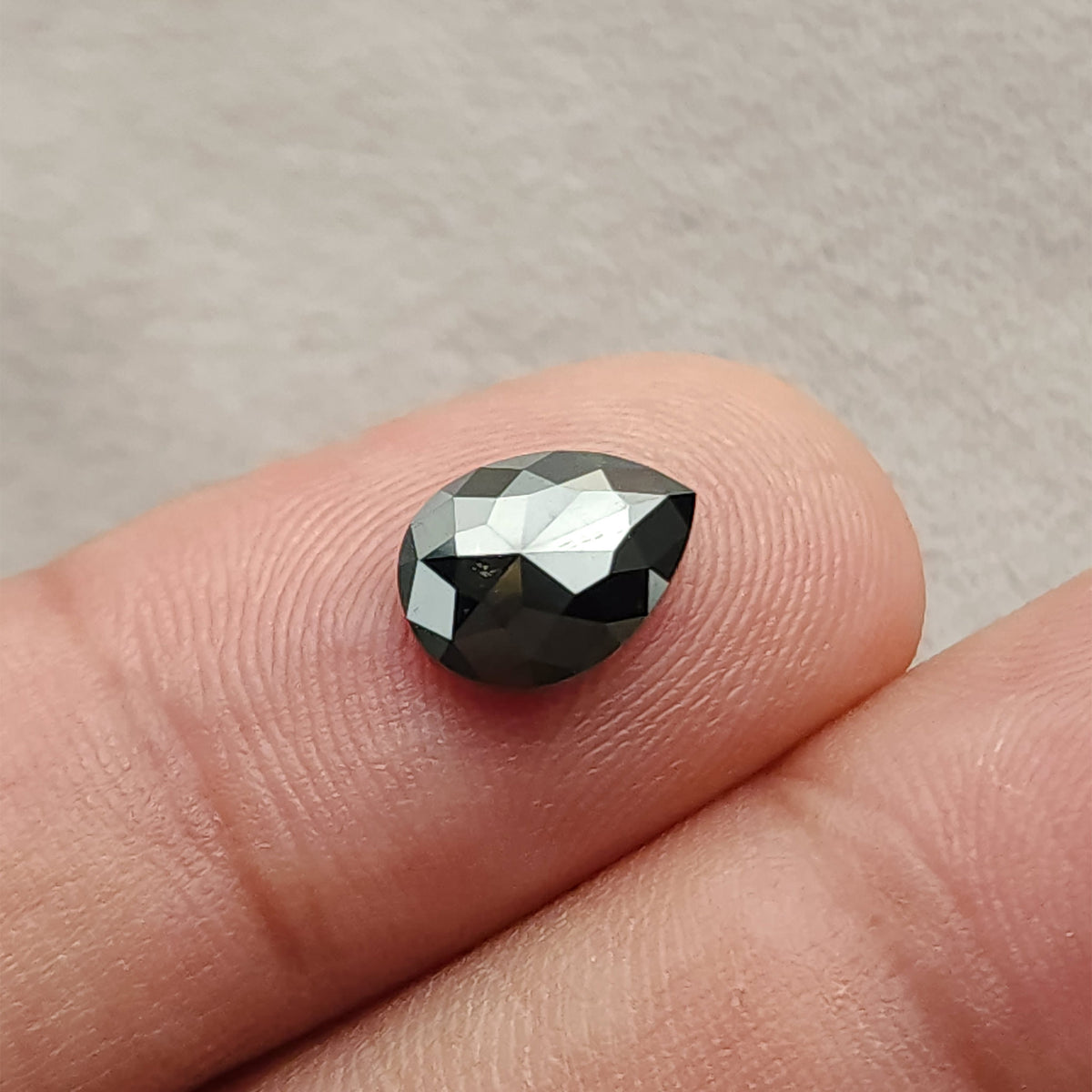 Low face-up angle of the opaque black diamond on a fingertip, showing its pear outline and bright reflections across the upper facets.