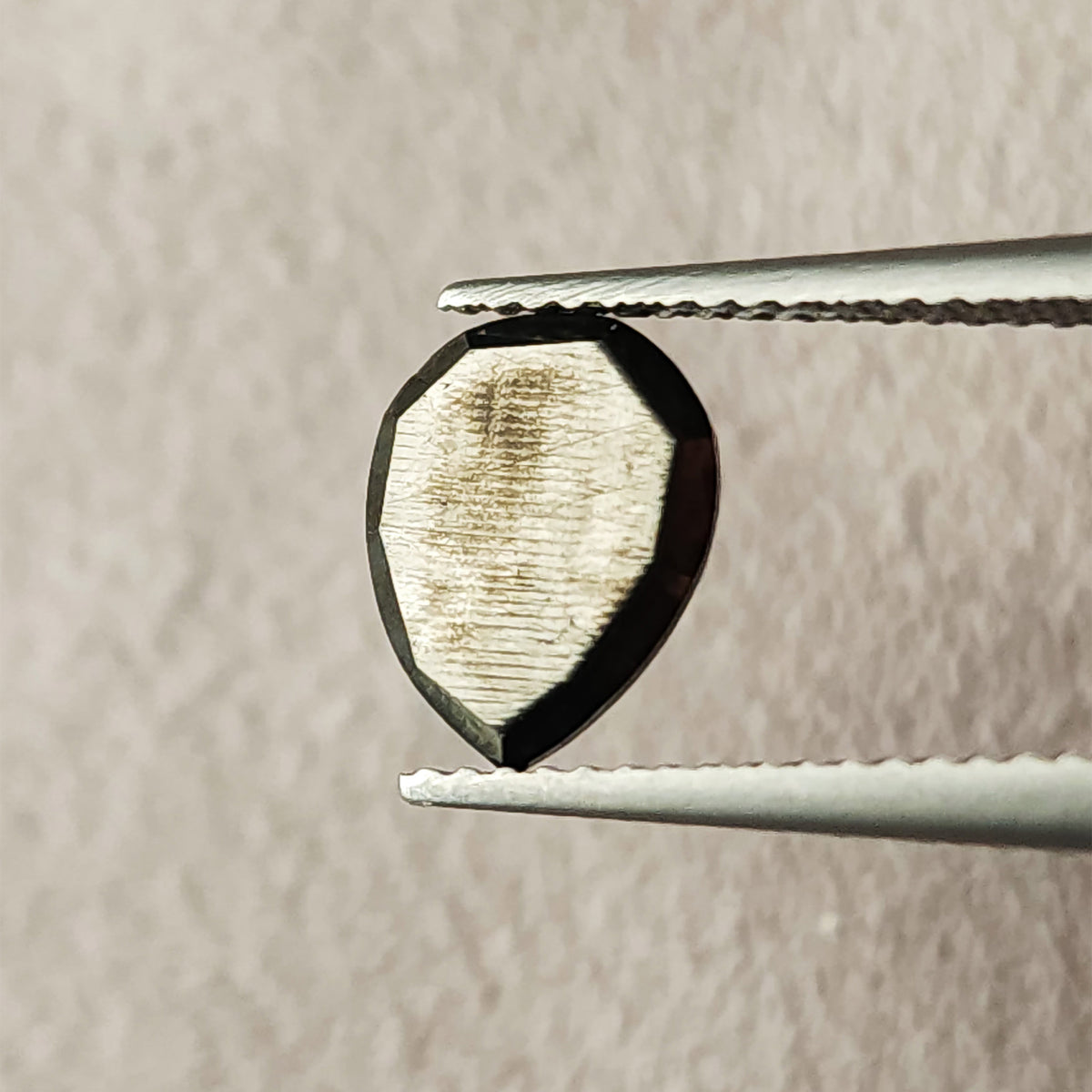 Reverse view of the pear rose-cut black diamond between tweezers, showing its flat back, pear outline and reflective surface.