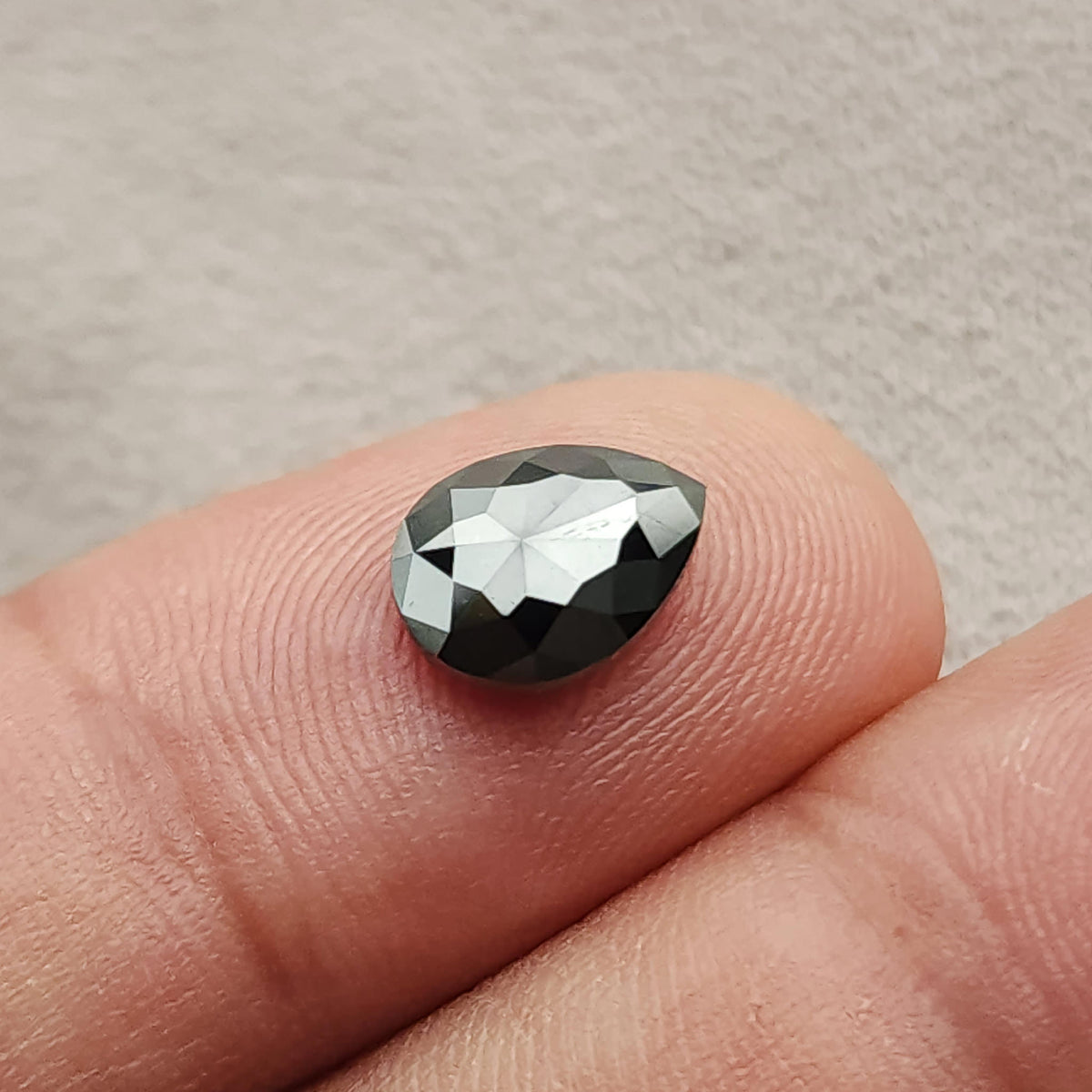 Alternate tilted view of the pear rose-cut black diamond on a fingertip, with broad mirror-like reflections across the center and darker outer facets.