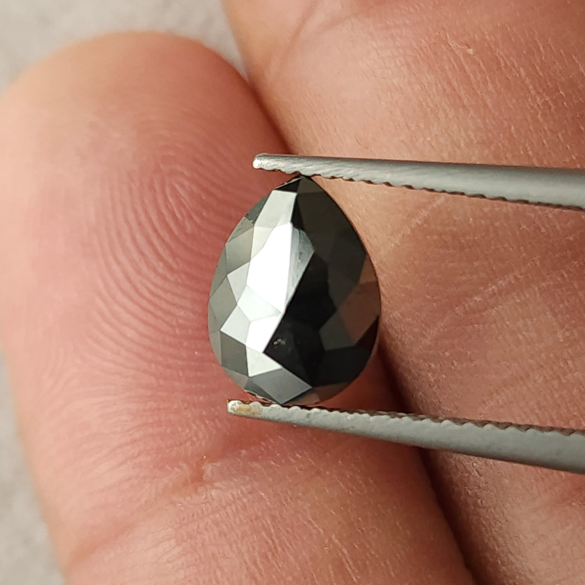 Face-up close-up of the black diamond between tweezers against fingers, providing real-world scale and showing alternating dark and bright rose-cut facets.
