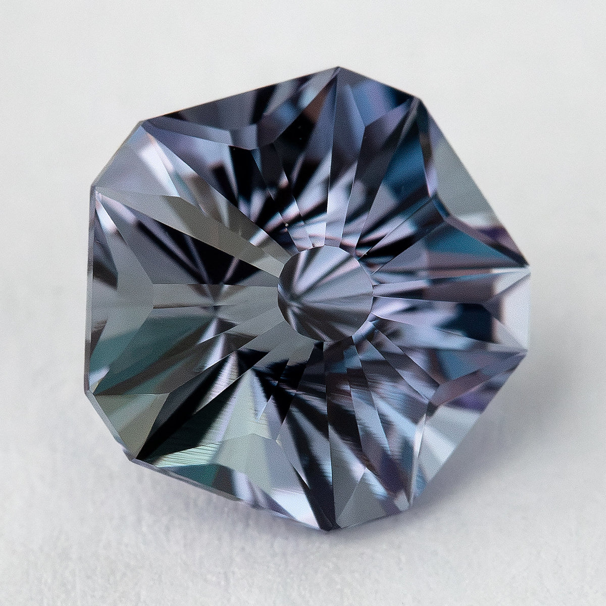 Angled Hanami-cut sapphire on a pale surface, with blue-gray, lavender and dark reflections across the crown.