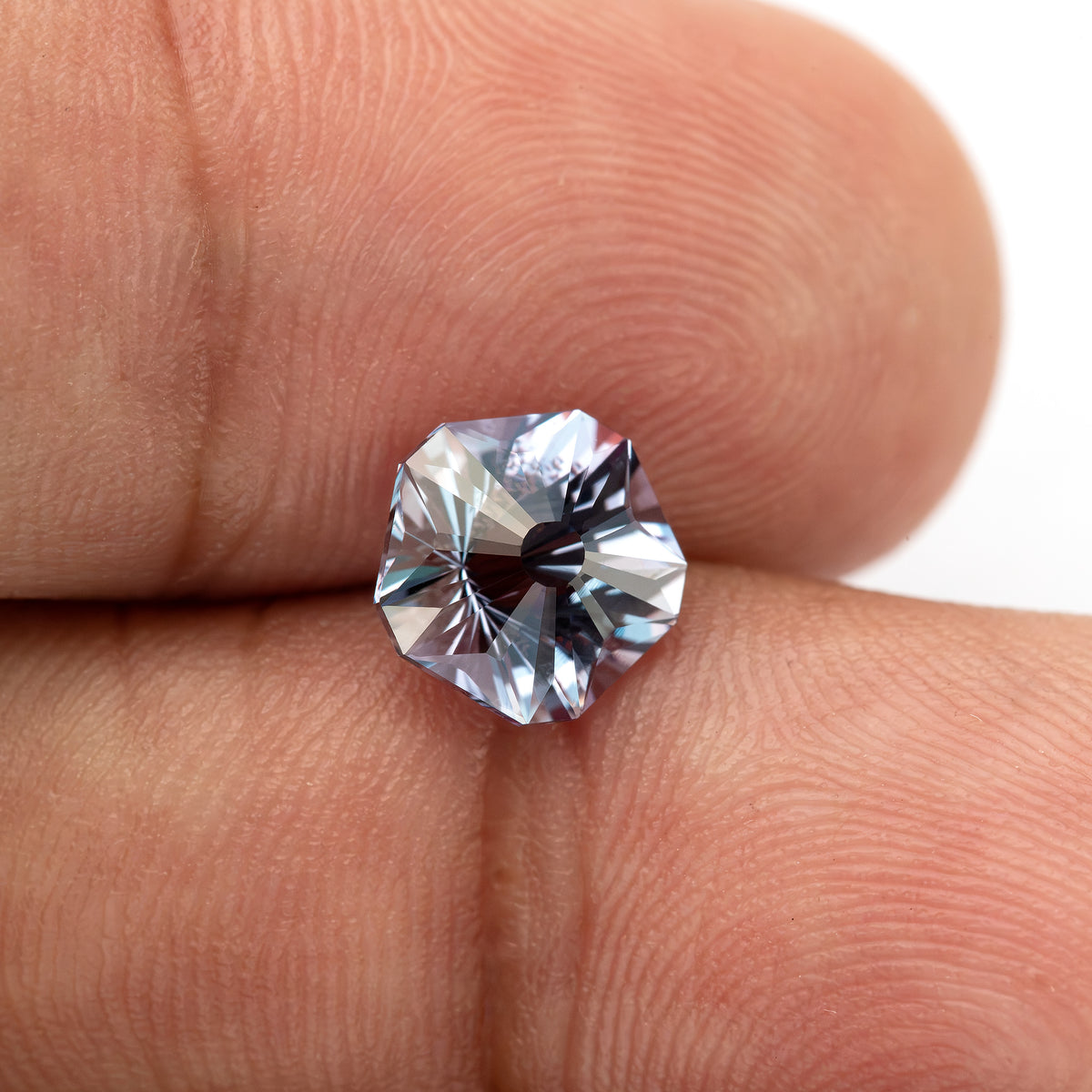 Face-up Hanami-cut sapphire resting between fingertips, with skin ridges showing its scale.