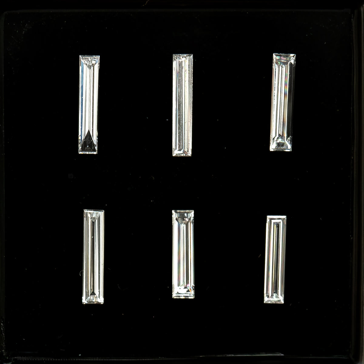 Six long baguette diamonds arranged in two rows of three on black backing, showing their different lengths, widths and step-facet reflections; sold individually by selection.