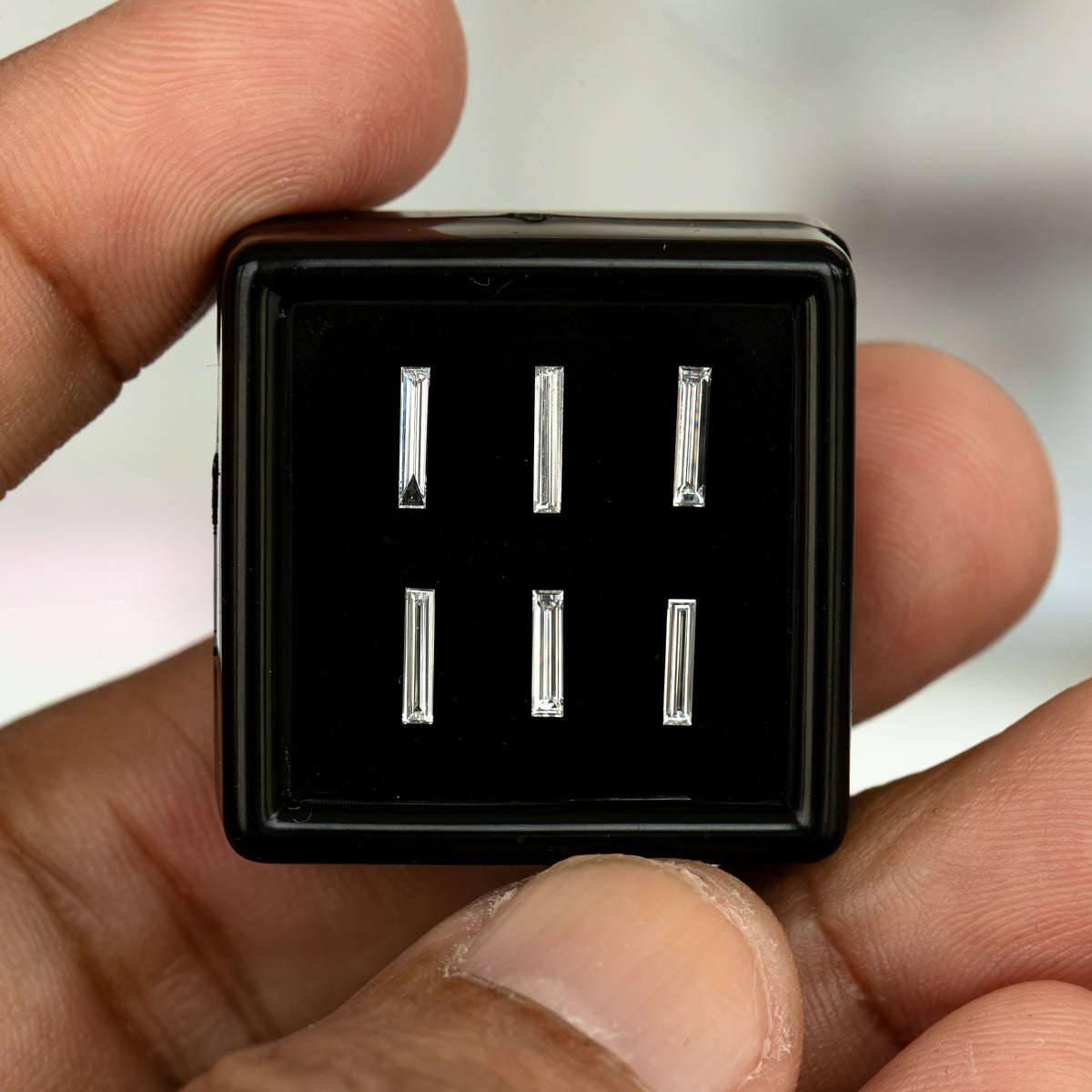 Black display box of six baguette-diamond choices held between fingers, showing the scale of the available individual options.