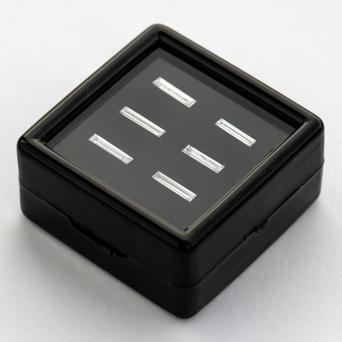 Low angled view of the black baguette-diamond display box on a pale surface, showing the tray depth and reflected faces of the six choices.