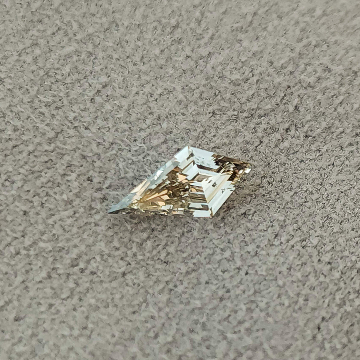 Angled view of the elongated kite lab-grown diamond resting on a textured gray surface, showing its outline and pale reflections.