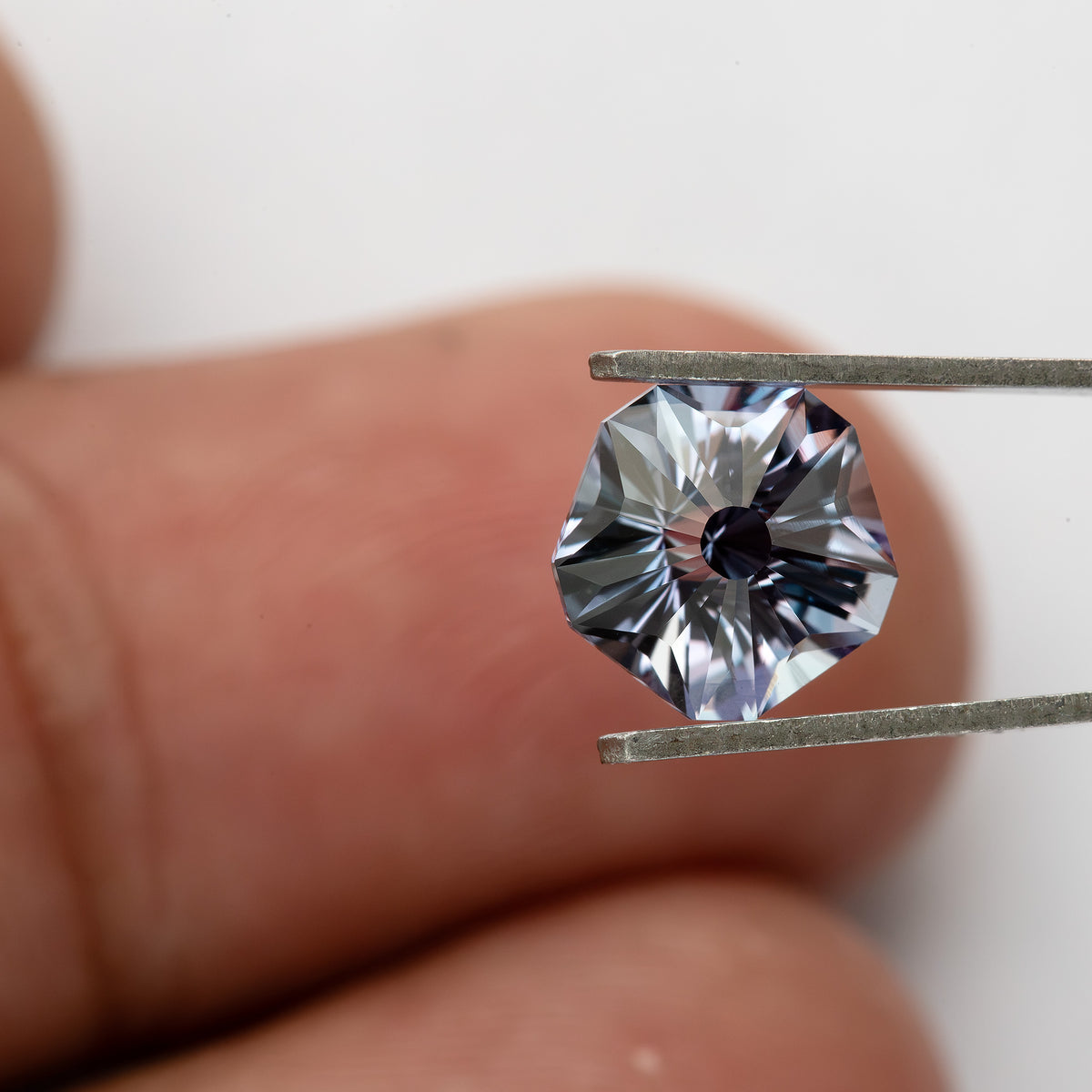 Single Hanami-cut sapphire held face-up in tweezers in front of fingertips for a scale reference.
