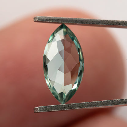 Mint-green marquise lab-grown sapphire held vertically between tweezers against a finger for scale.