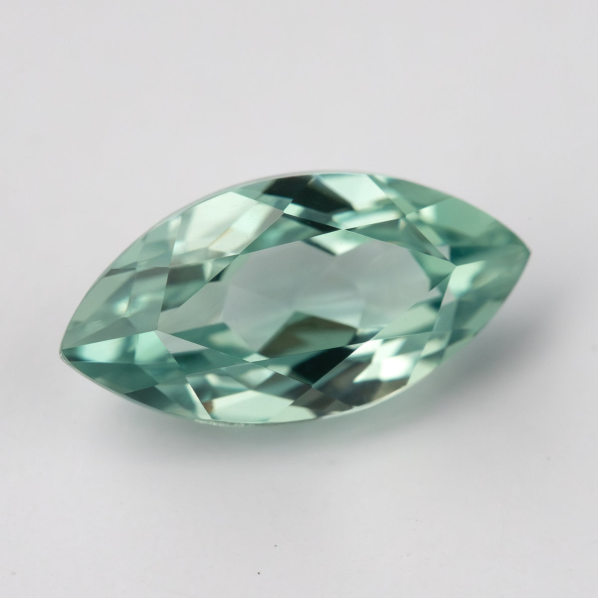 Tilted view of a mint-green marquise lab-grown sapphire on a pale surface, showing green facet reflections.