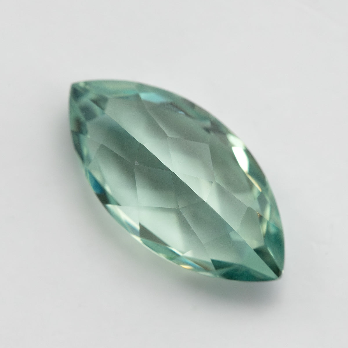 Diagonal view of a mint-green marquise lab-grown sapphire on a pale surface, showing pale green facet reflections.