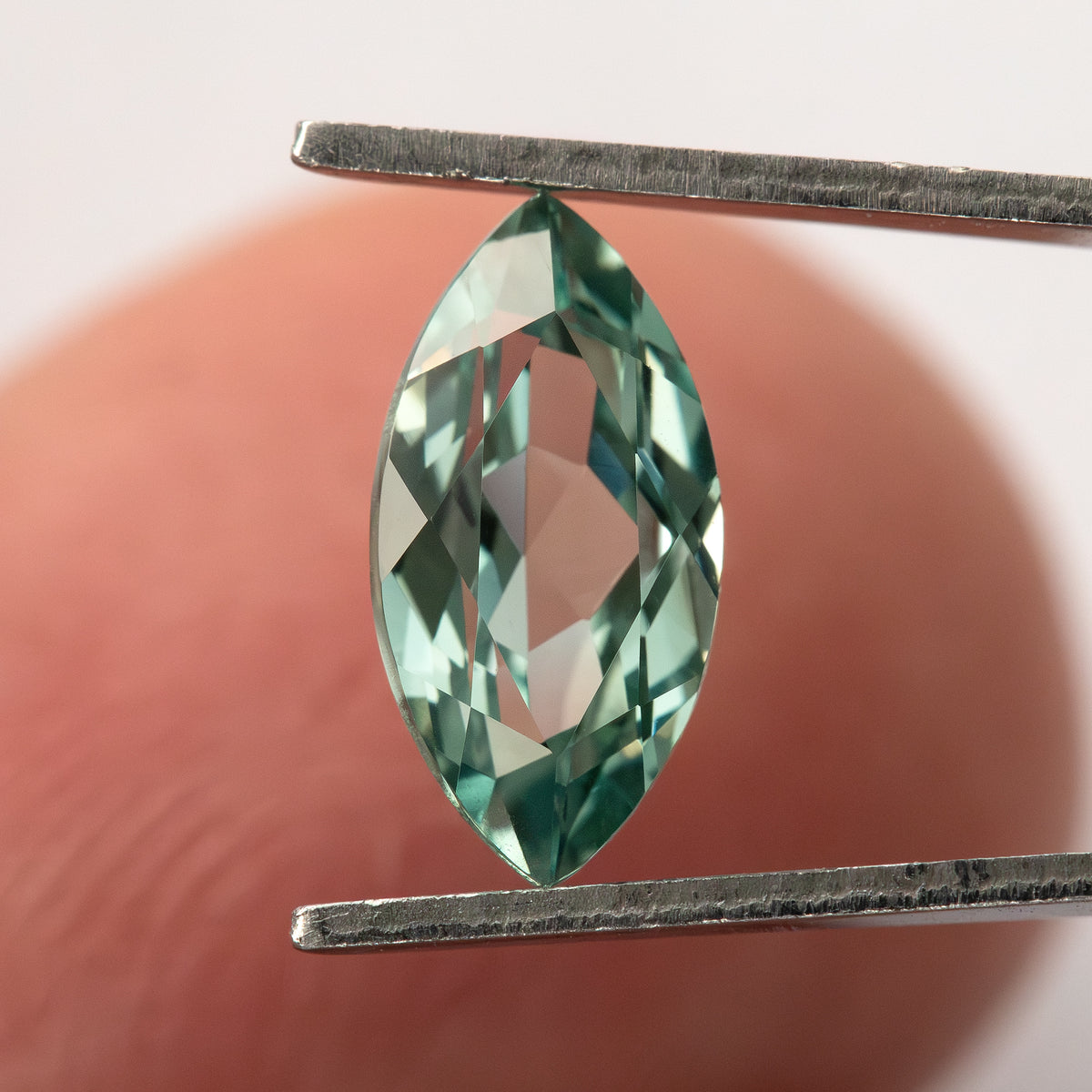 Mint-green marquise lab-grown sapphire held upright between tweezers against a fingertip, showing its size and faceted outline.