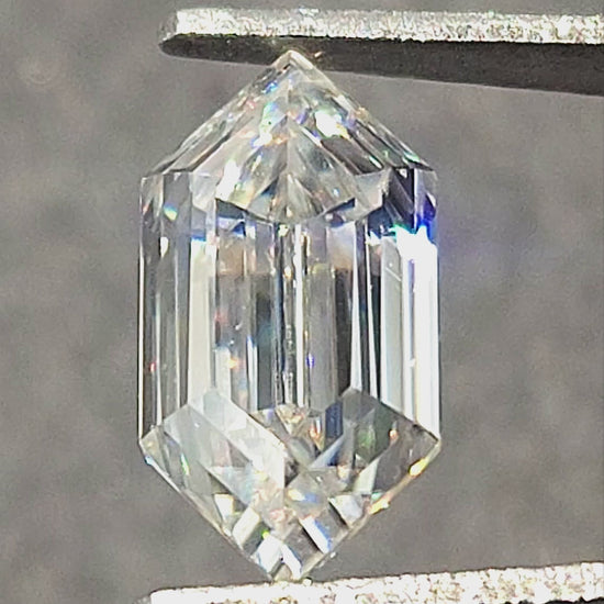 Elongated hexagon step-cut moissanite rotated in tweezers and resting on fingertips, showing facet reflections and rainbow flashes.