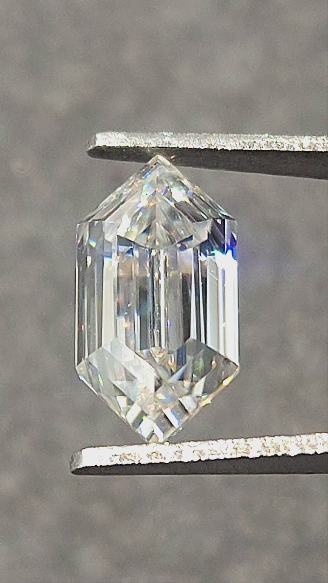 Elongated hexagon step-cut moissanite rotated in tweezers and resting on fingertips, showing facet reflections and rainbow flashes.
