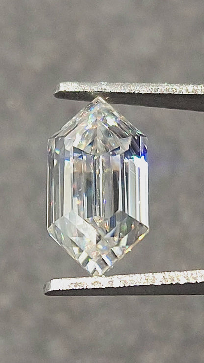Elongated hexagon step-cut moissanite rotated in tweezers and resting on fingertips, showing facet reflections and rainbow flashes.