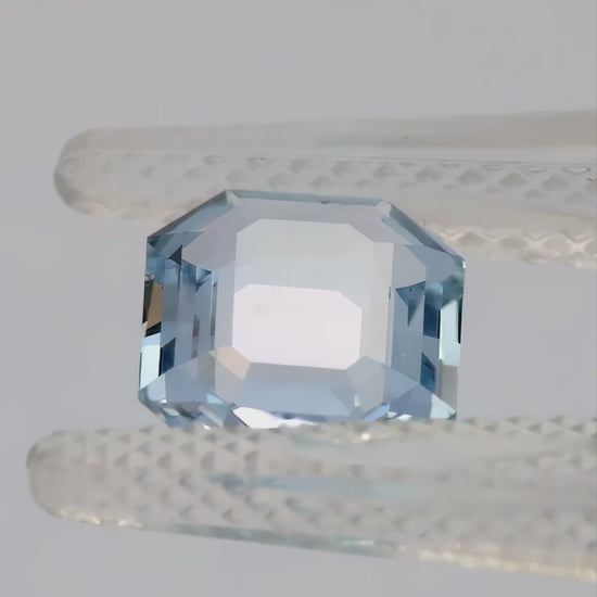 Light blue portrait-cut octagonal sapphire tilted in tweezers and resting face-up on a fingertip.