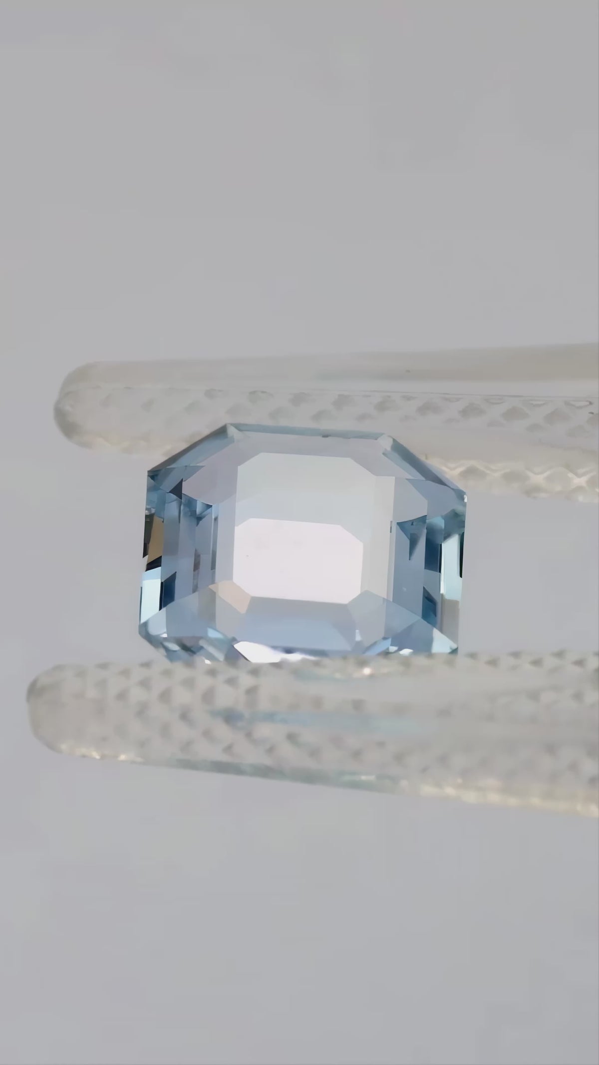 Light blue portrait-cut octagonal sapphire tilted in tweezers and resting face-up on a fingertip.