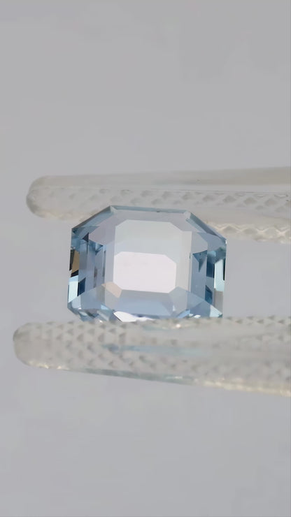 Light blue portrait-cut octagonal sapphire tilted in tweezers and resting face-up on a fingertip.